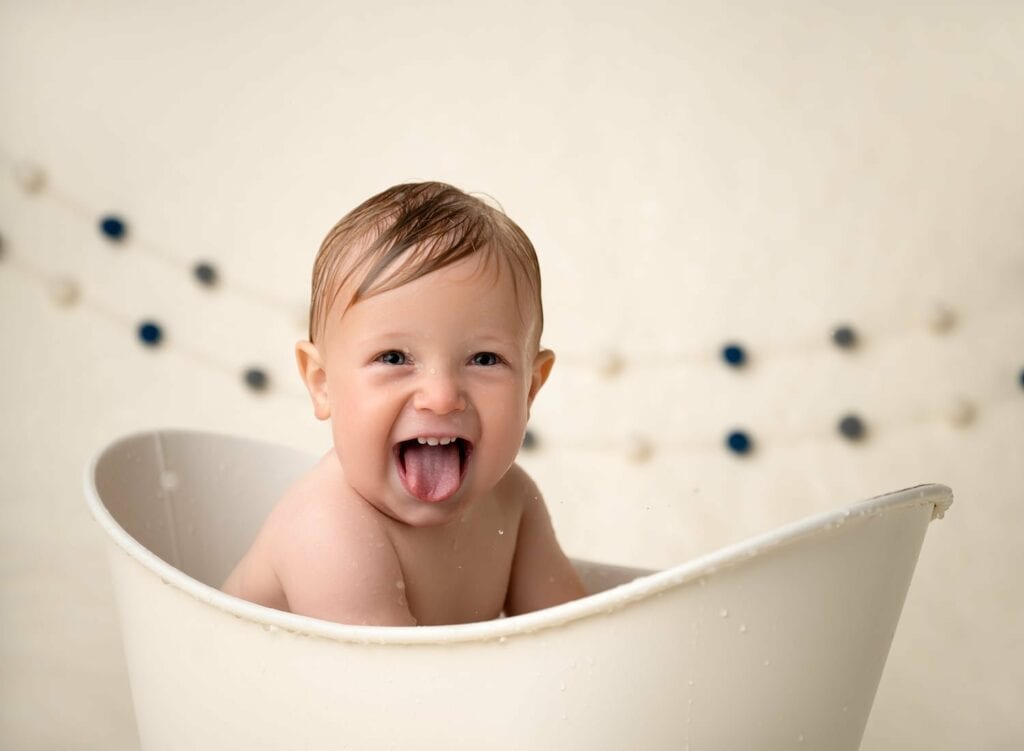 Baby sitting in a prop bathtub, and sticking out his tongue while smiling. 