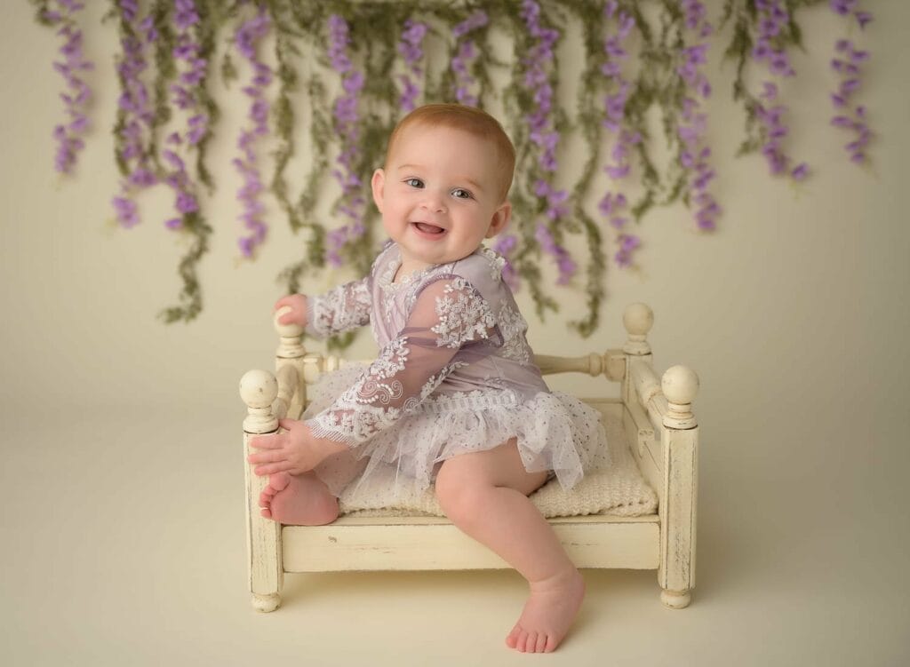 Baby girl sitting on a small vintage couch with lavender in the background.