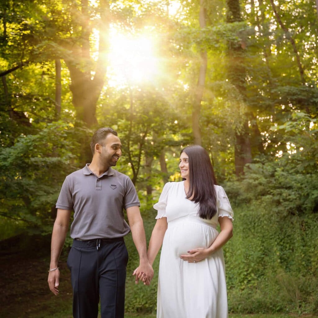 A couple holding hands in front of trees with the sun shining through. The woman is holding her belly to show she is pregnant.